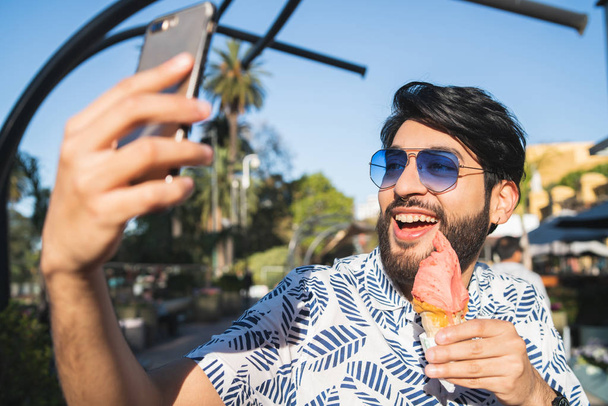 Portrait of young man enjoying sunny weather, taking a selfie with the phone and eating an ice cream outdoors. - Φωτογραφία, εικόνα