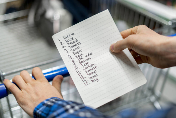 manos de una persona sosteniendo una hoja de papel de la lista de la compra y comprobar la compra de productos en la tienda de comestibles
 - Foto, Imagen