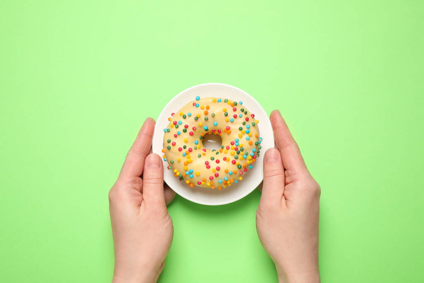 Woman holding plate with delicious glazed donut on green background, top view - Photo, Image
