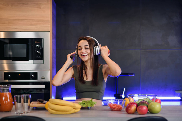 Mujer atractiva joven come manzana roja y escucha la música en la cocina por la mañana. Concepto de estilo de vida saludable, disfrutar del desayuno
. - Foto, imagen