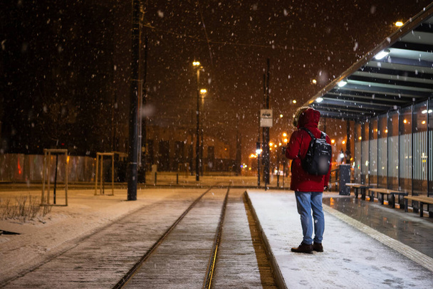 「赤い冬のコートを着た男がバス停に立って公共交通機関を待ってる」 - 写真・画像