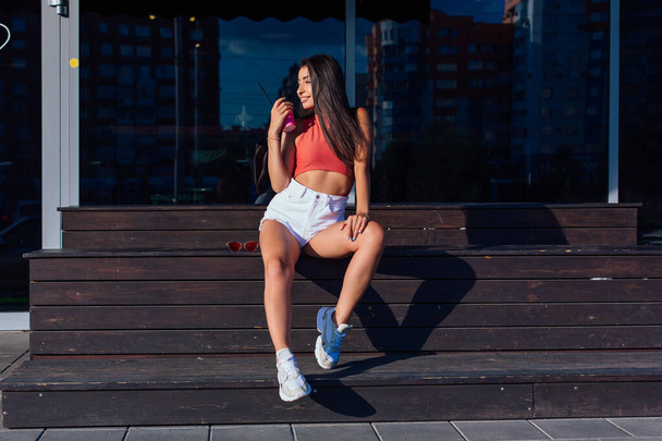 Stylish happy young woman wearing white shorts and sneakers holding pink cup of coffee to go sitting next to coffee shop. Portrait of smiling girl sitting on the bench with coffee. - 写真・画像