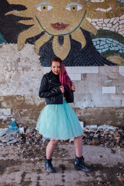 Young girl with pink hair dressed in leather jacket and a blue skirt standing inside of collapsed building surrounded by ruins. Woman standing next to the old mosaic wall on the background. - Fotografie, Obrázek