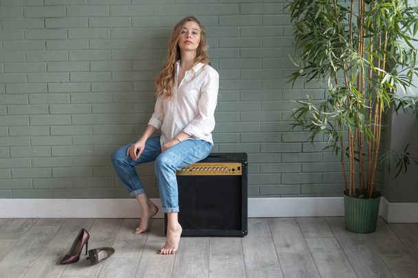 Young beautiful blond woman wearing white shirt and blue jeans sitting on subwoofer with bare feet next to green plant. Girl sitting in a daylight from the window. Copy space - 写真・画像