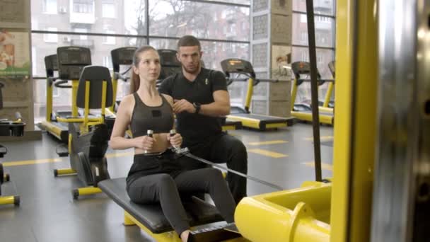 Mujer joven con instructor haciendo ejercicio en el gimnasio mediante el uso de la máquina de remo
 - Metraje, vídeo