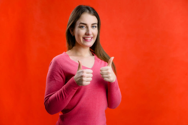 Portrait On a red background of a pretty smiling young woman in a pink sweater shows hands ok - Фото, зображення