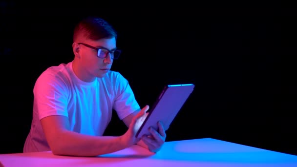 Un joven con una tableta. Un hombre está usando una tableta. Luz azul y roja cae sobre un hombre sobre un fondo negro
. - Imágenes, Vídeo