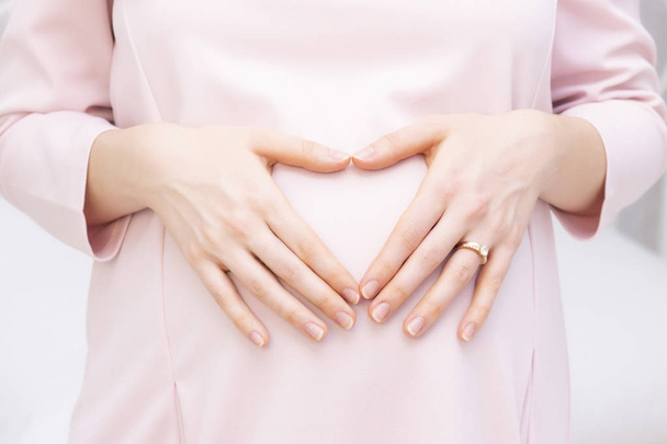 Een zwangere vrouw in een jurk houdt haar handen op haar buik in de vorm van een hart. Het concept van zwangerschap, moederschap, voorbereiding en verwachting. Close-up binnen. - Foto, afbeelding