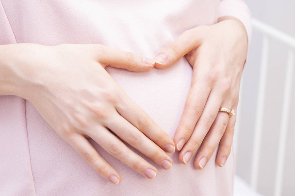 pregnant woman in a dress holds her hands on her stomach in the shape of a heart. The concept of pregnancy, motherhood, preparation and expectation. Close-up indoors. - Photo, Image
