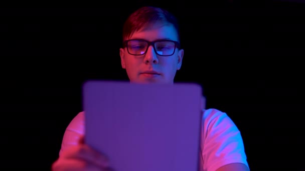 Un giovanotto con un tablet. Un uomo usa una tavoletta. Luce blu e rossa cade su un uomo su uno sfondo nero
. - Filmati, video