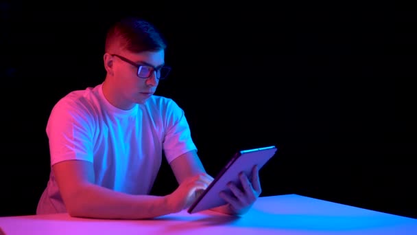 Un joven con una tableta. Un hombre está usando una tableta. Luz azul y roja cae sobre un hombre sobre un fondo negro
. - Imágenes, Vídeo