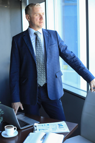 Mature businessman in a corporate suit standing in office and looking away through large windows optimistically - Zdjęcie, obraz