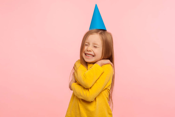 Fascinating lovely cute little girl with funny party cone on head embracing herself and keeping eyes closed, self-love concept, positive self-esteem. indoor studio shot isolated on pink background - Photo, Image