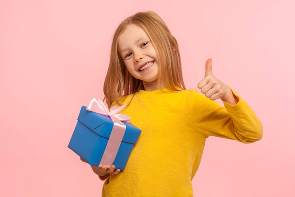 Best present! Portrait of lovely happy smiling little girl holding wrapped box and showing thumbs up, like gesture, child satisfied with birthday gift. indoor studio shot isolated on pink background - Fotoğraf, Görsel