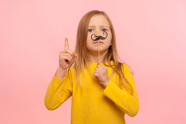 Bossy little ginger girl with funny paper mustaches gesturing finer up, giving advice, pretending to be smart male, having fun with masquerade accessory. indoor studio shot isolated on pink background - 写真・画像