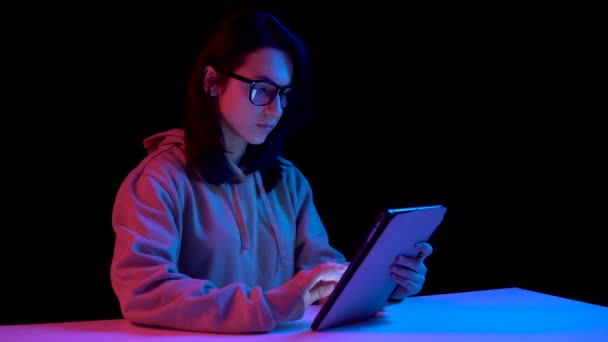 Mujer joven con una tableta. Una mujer está usando una tableta. Luz azul y roja cae sobre una mujer sobre un fondo negro
. - Imágenes, Vídeo
