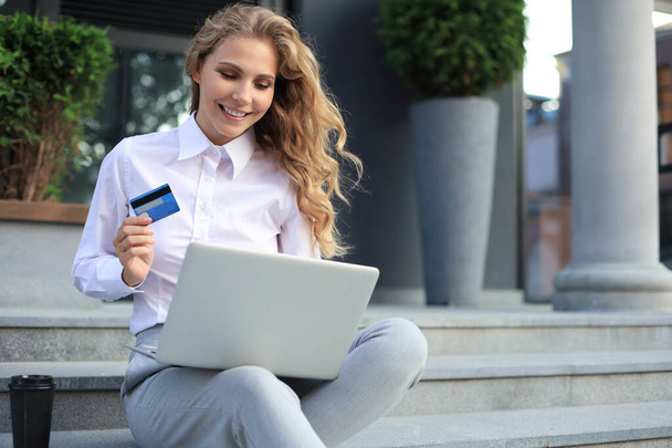 Online shopping concept. Young blonde woman holding a credit card and doing online payment with laptop outdoors - Foto, afbeelding