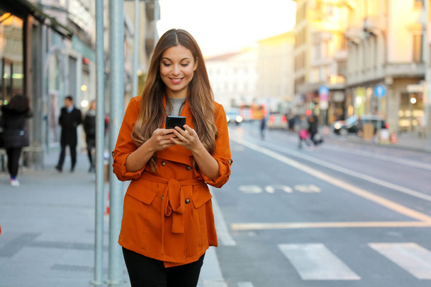 Portrait of beautiful smiling woman walking in city street texting on mobile phone with blurred background - Photo, Image