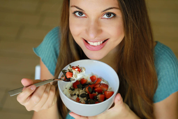 Close-up de cima de bela jovem mulher sorrindo e comendo skyr com frutas de muesli de cereais e sementes em casa, foco nos olhos modelo, imagem interior. Conceito de alimentação saudável
. - Foto, Imagem