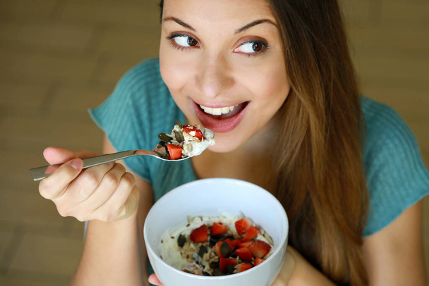 Вид сверху на красивую молодую женщину, которая ест йогурт с клубникой, зерновыми мюсли и семенами дома, смотрит в сторону, фокусируется на образцовых глазах, крытый снимок. Концепция здорового питания
. - Фото, изображение