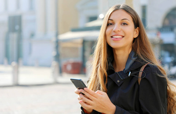 Femme heureuse souriant et marchant dans la rue à l'aide d'un smartphone et regardant la caméra - Photo, image