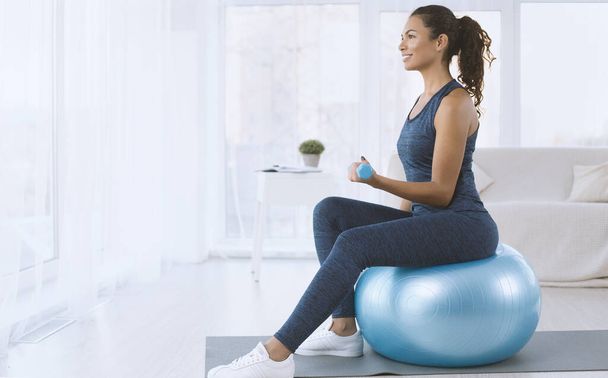 Senhora hispânica atraente com halteres sentados na bola de fitness em casa, espaço de cópia
 - Foto, Imagem