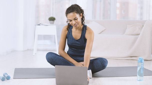 Hispanic woman with laptop watching workout video on yoga mat indoors - Foto, Bild