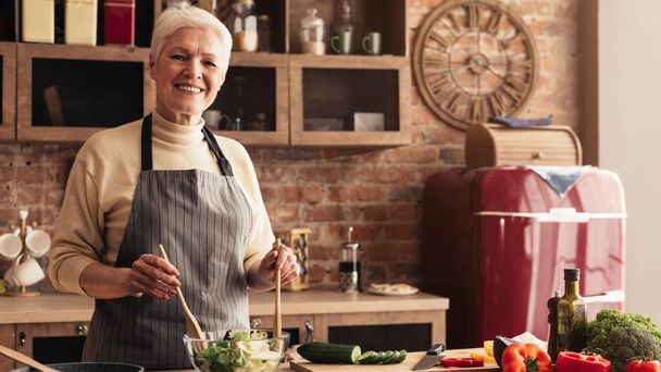 Retrato de mulher idosa sorrindo cozinhar salada de legumes frescos na cozinha
 - Foto, Imagem
