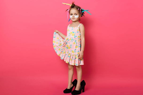Horizontal shot of little girl wearing big mothers shoes and sundress, posing with hair curlers, cute kid standing with patches under eyes isolated over rose background. Child fashion concept. - 写真・画像