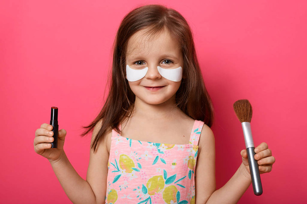 Close up portrait of cute little girl with brush and pomade in hands, doing make up with mother's cosmetic, has patches under her eyes, looks smiling at camera, wears sundress, stands over pink wall. - Foto, Imagem