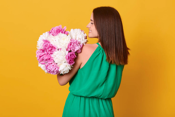 Back view of charming stylish woman wearing elegant green sundress isolated over yellow studio background, holding big peony flower bouquet, lady with dark srtraight hair, has present from husband. - Foto, imagen