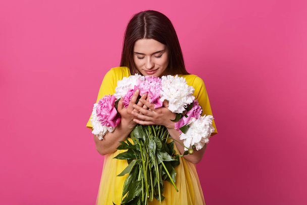 Close up portrait of beautiful brunette woman holding flowers isolated over pink studio background, lady wearing yellow dress embracing big bouquet, girl looking at flowers. 8 march concept. - 写真・画像