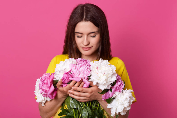 Indoor shot of beautiful dark haired female embracing big bouquet with pink and white peony flowers, has gentle look, posing isolated over pink studio background. Women's Day, 8 march concept. - Photo, image