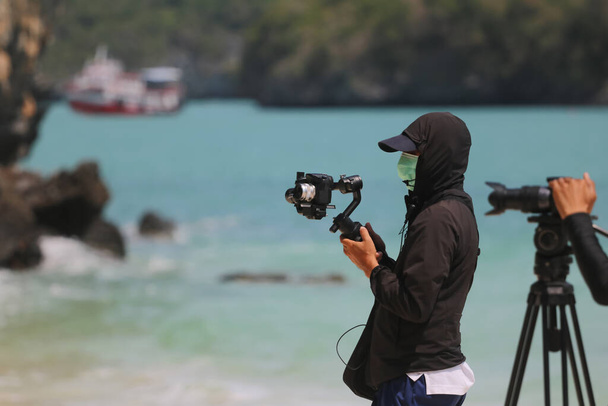 マスクを着用し、コロナウイルスやcovid -19の普及中にビーチでの観光活動を記録したビデオ写真家やジャーナリスト. - 写真・画像