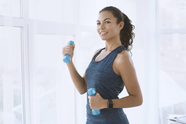 Happy Hispanic woman with dumbbells on her workout indoors, space for text - Photo, Image
