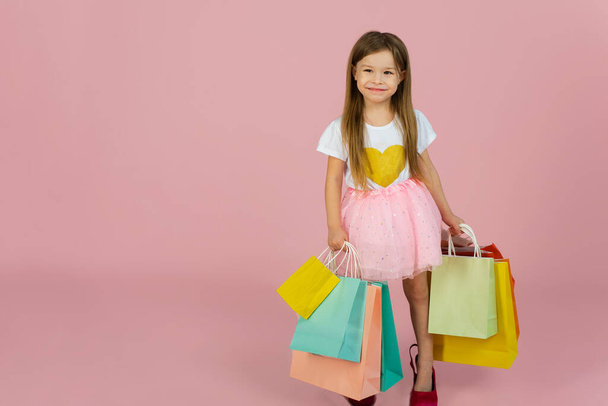 Dívka s mnoha nákupních tašek na pastelově růžový backgound. Krásné sladké okamžiky malé princezny, docela přátelské dítě baví na kameru. - Fotografie, Obrázek