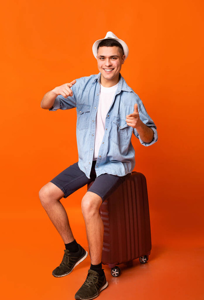 Счастливый молодой турист сидит на чемодане и указывает на камеру
 - Фото, изображение