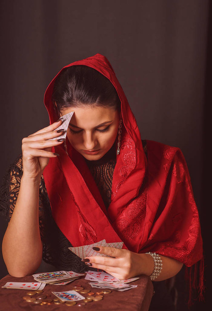Mujer con una bufanda en la cabeza y con cartas en las manos adivina sobre una vela y monedas
 - Foto, imagen