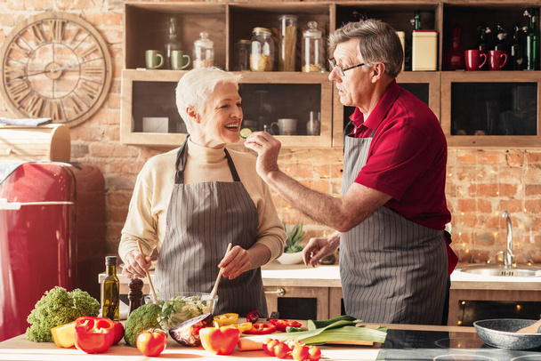 Romantic aged couple tasting food while cooking lunch in kitchen together - Zdjęcie, obraz