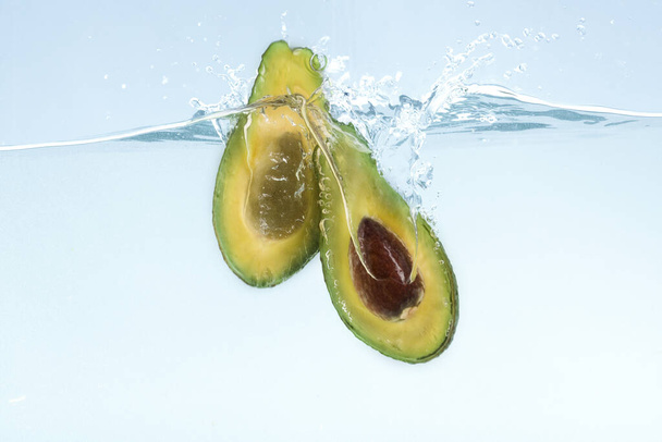 Tasty avocado cut in half dropping into transparent water on blue background - Foto, Imagen