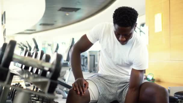 Young African American man sitting and lifting a dumbbell close to the rack at gym. Male weight training person doing a biceps curl in fitness center - Video