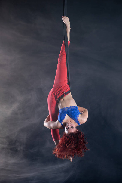 Female athletic, sexy and flexible aerial circus artist with redhead on aerial straps on black background - Фото, изображение
