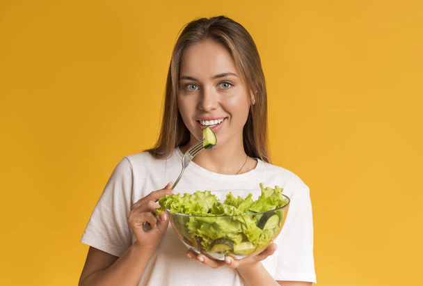 Счастливая молодая женщина ест свежий овощной салат, позируя на желтом фоне
 - Фото, изображение