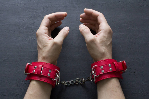 赤いエロ革の手錠で大人の男性の手。監禁されてる。黒の背景に赤い革の手錠。大人のゲームやおもちゃ。BDSM,フェチウェアとキンキーなセックスおもちゃのコンセプトとともにクローズアップ. - 写真・画像