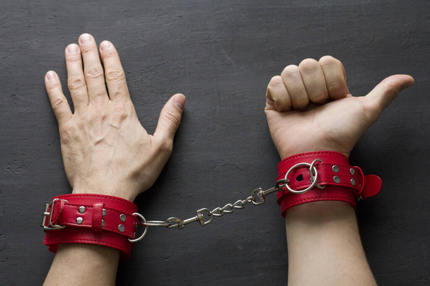 Adult male hands in red erotic leather handcuffs. Hands in captivity. Red leather handcuffs on black background. Adult games and toys. BDSM, fetish wear and kinky sex toy concept with close up. - Фото, зображення