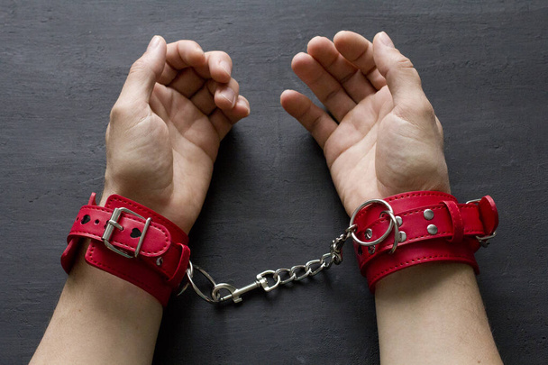 Adult male hands in red erotic leather handcuffs. Hands in captivity. Red leather handcuffs on black background. Adult games and toys. BDSM, fetish wear and kinky sex toy concept with close up. - Photo, Image