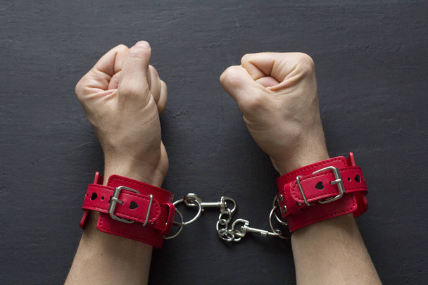 Adult male hands in red erotic leather handcuffs. Hands in captivity. Red leather handcuffs on black background. Adult games and toys. BDSM, fetish wear and kinky sex toy concept with close up. - Photo, Image