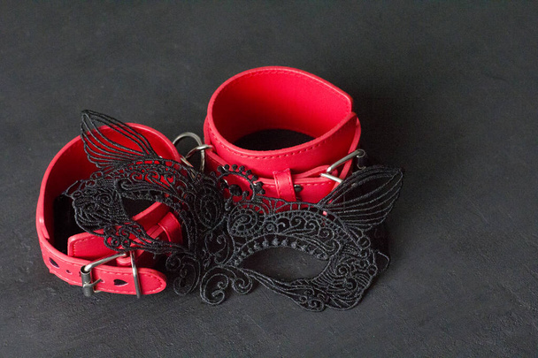 Красные кожаные наручники, эротическая черная маска на лице на черном фоне. Игры для взрослых и игрушки. БДСМ, бондаж, фетиш-одежда и извращенная концепция секс-игрушек с крупным планом
. - Фото, изображение