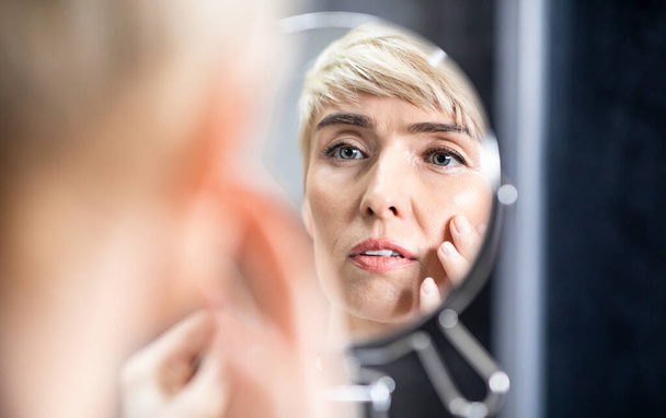 Зріла жінка дивлячись у дзеркало торкаючись обличчя стоячи у ванній
 - Фото, зображення