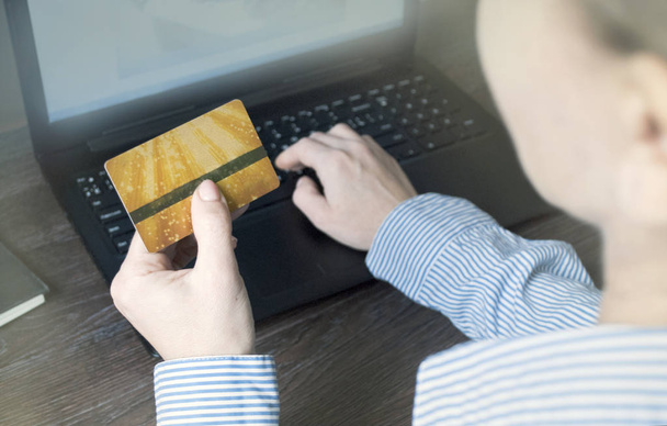 Kadın ellerinin kredi kartı tutarak ve bilgisayar kullanarak arkadan bakmasını. Çalışma alanı. Altın kredi kartı. - Fotoğraf, Görsel